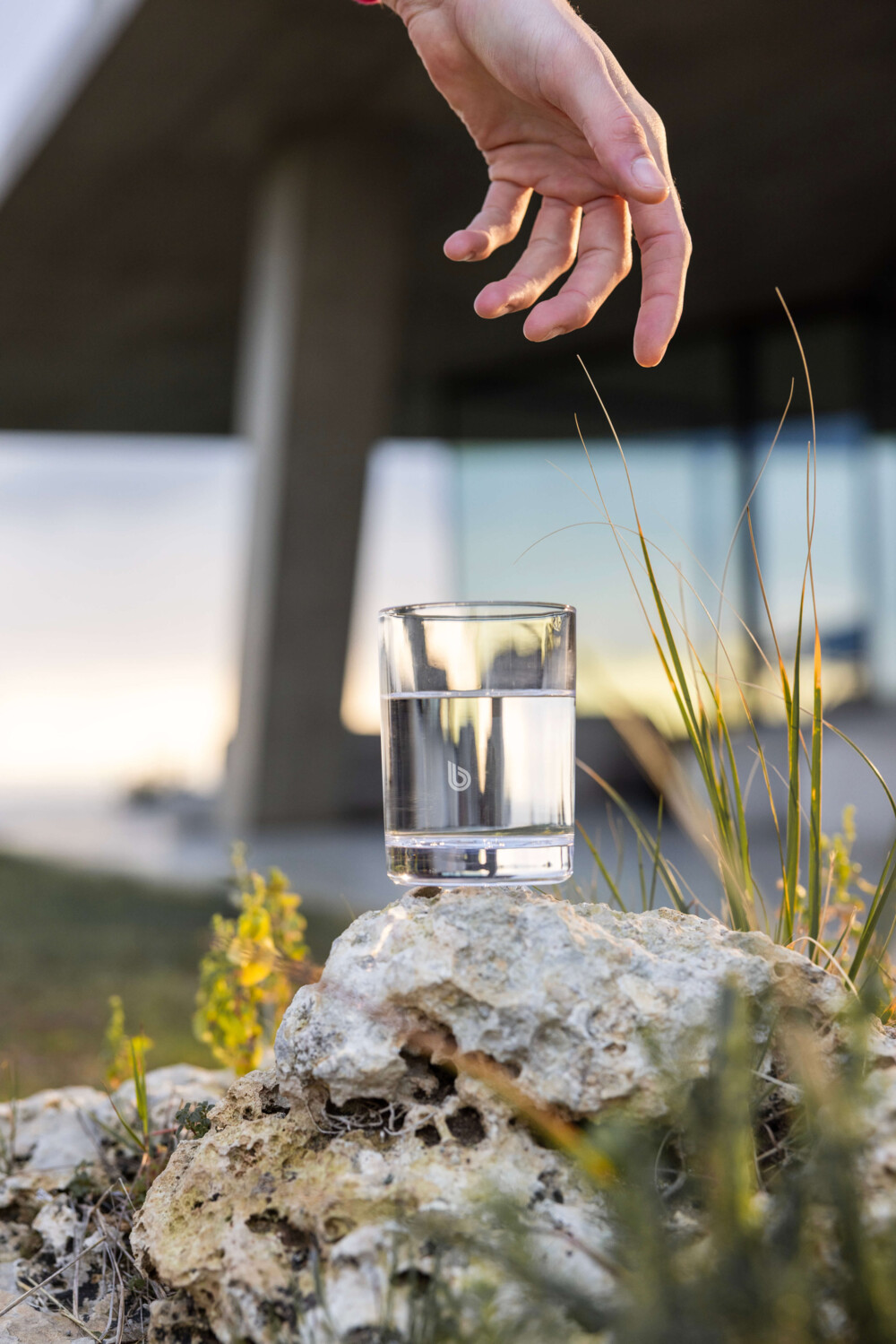 A clear glass of BWT pearlwater rests on a rocky surface surrounded by small plants, with a hand extending above as if to pick it up. 
